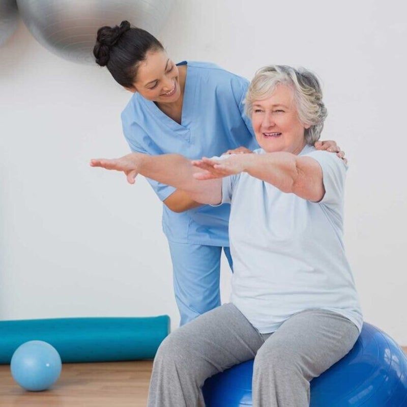 Una donna è seduta su una palla da ginnastica blu e si allunga con l'aiuto di un operatore sanitario in palestra.