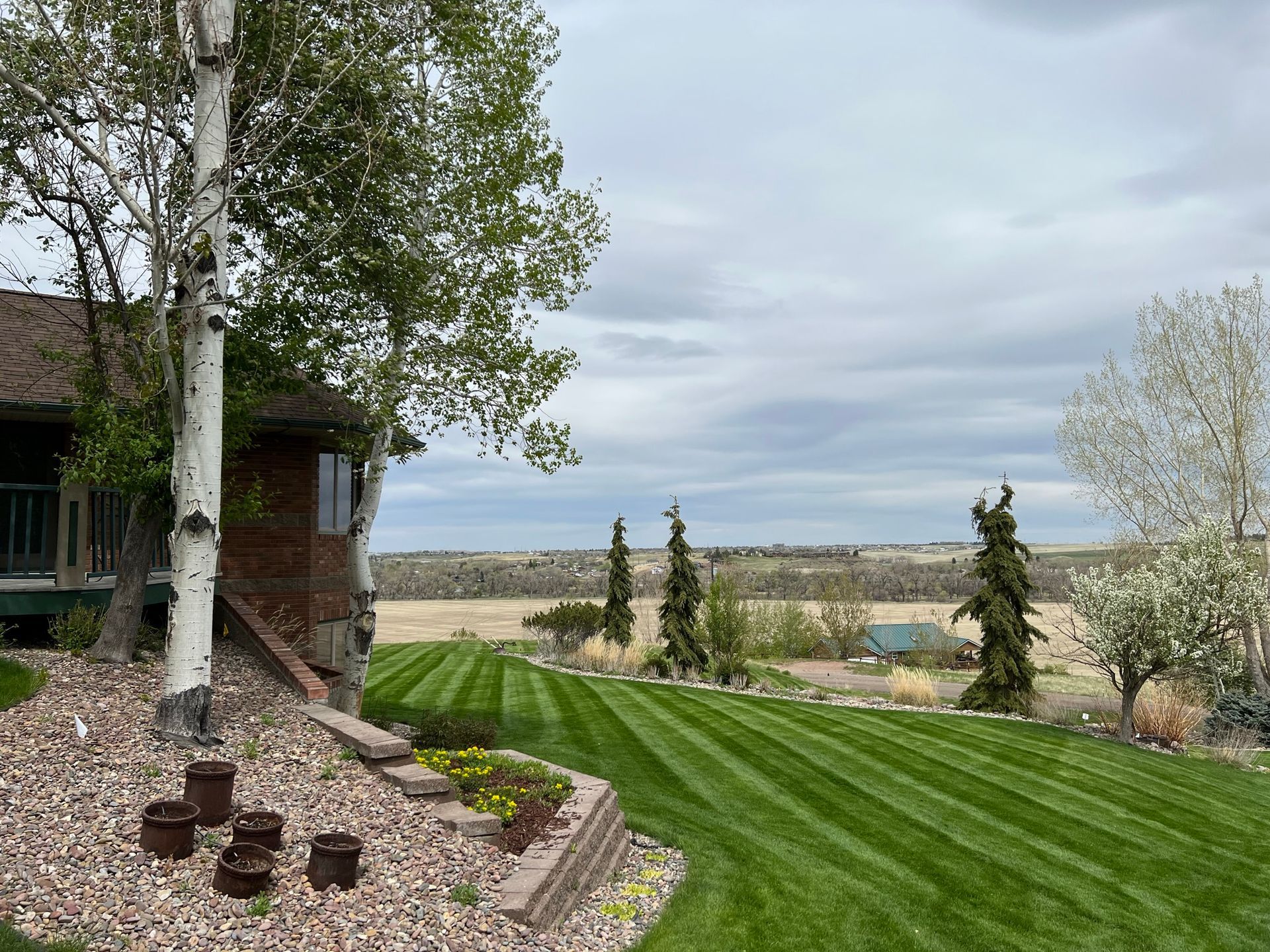 Lush green lawn with striped pattern, trees, and a house overlooking a distant landscape under a cloudy sky.