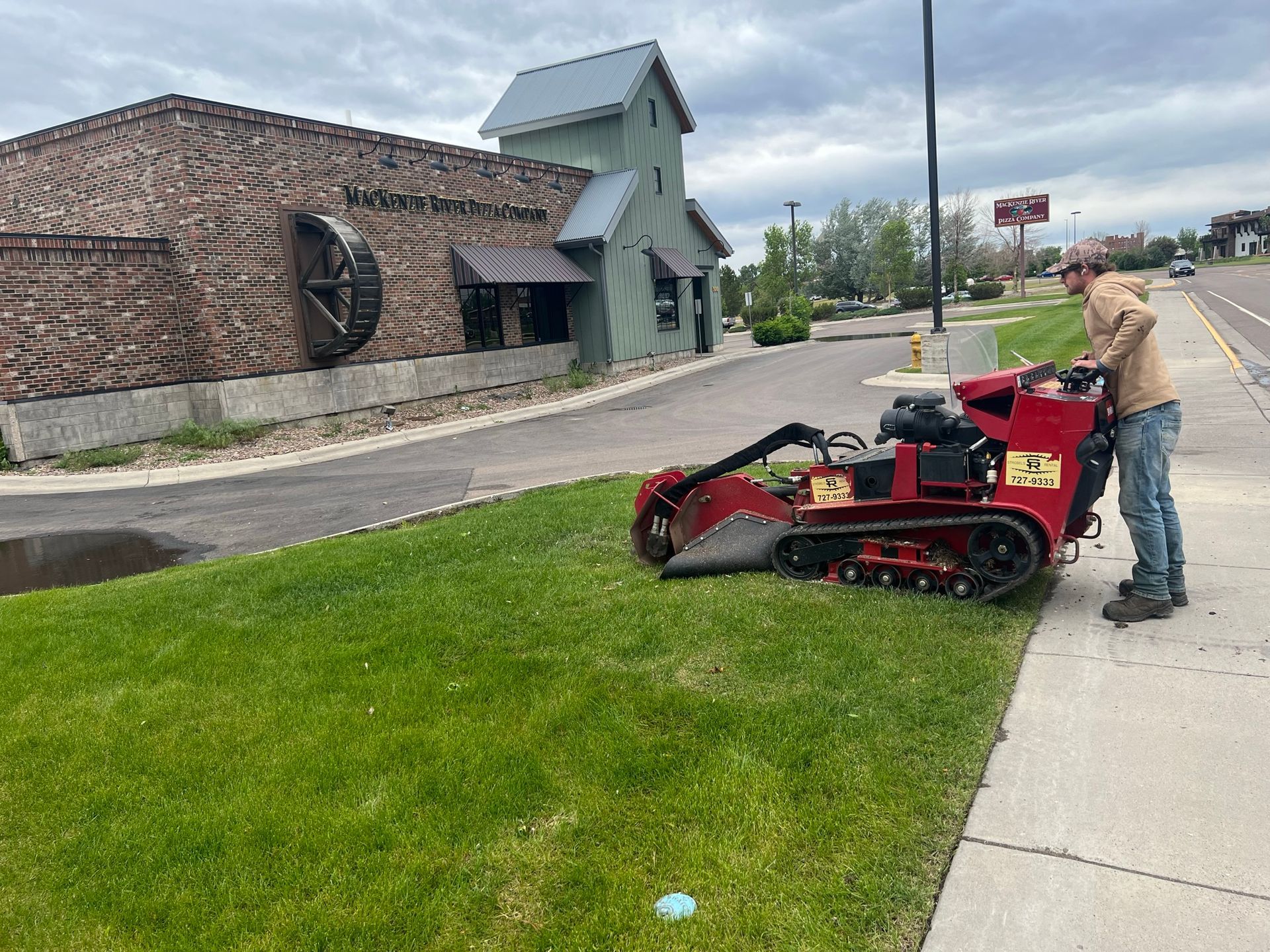 Man operating a stump grinder on a grassy area near a sidewalk and building with a brick exterior.