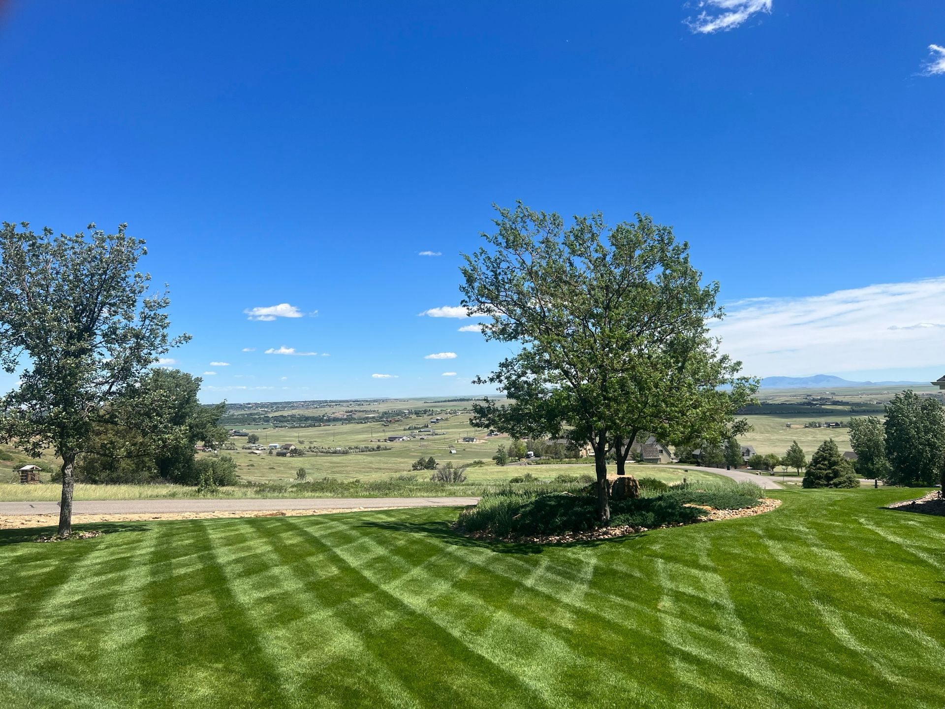 Lush green lawn with striped mowing pattern, trees, and a scenic valley under a bright blue sky.