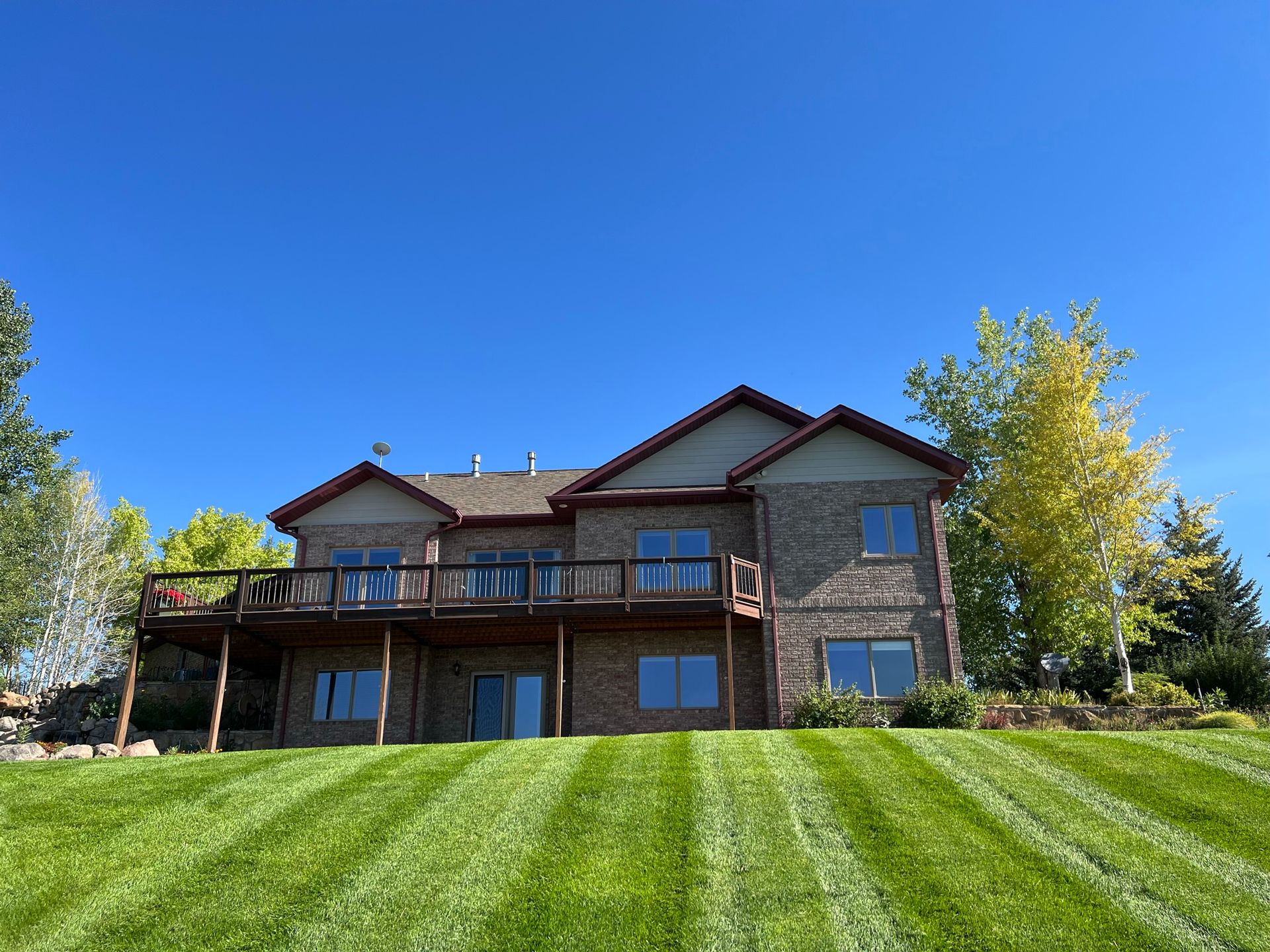Two-story brick house with a deck on a grassy hill under a bright blue sky.