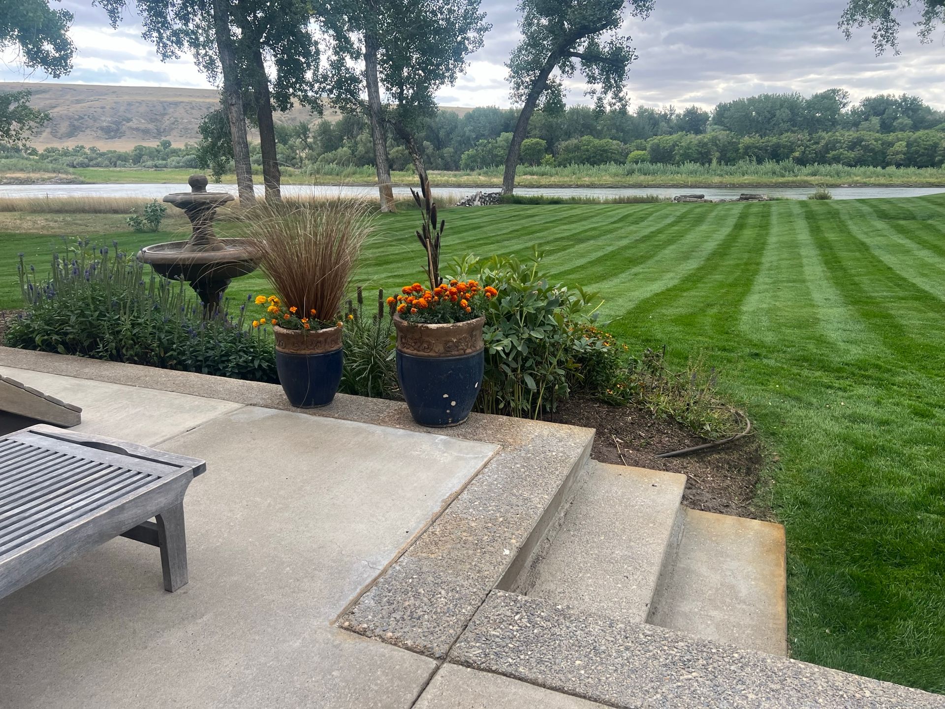 Patio with planters, fountain, and striped lawn leading to a river. Overcast sky.