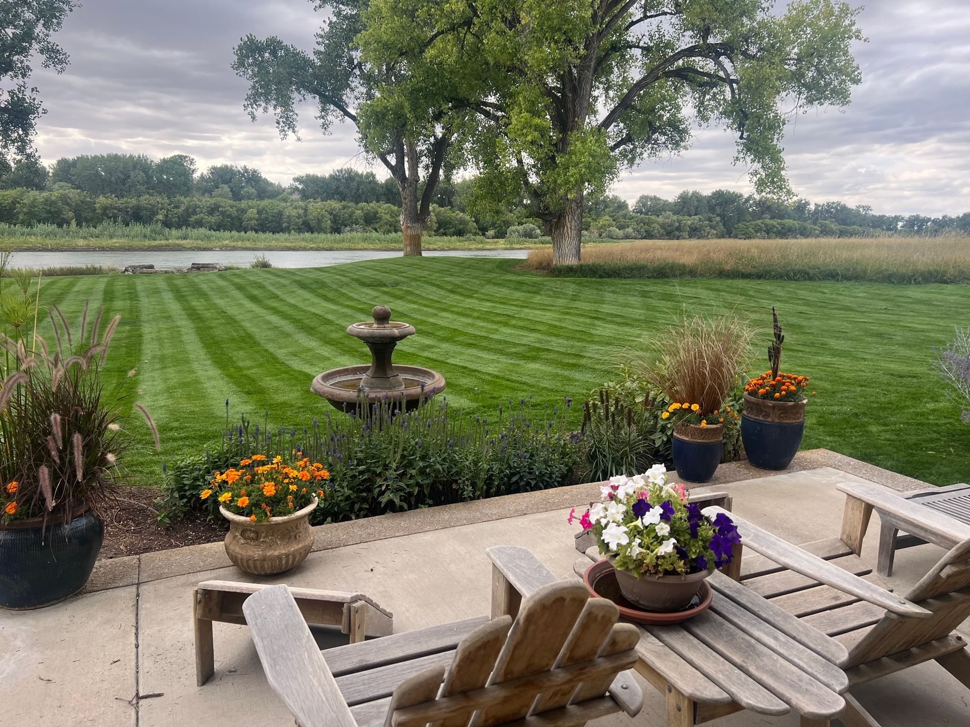 Lawn with fountain, potted flowers, and adirondack chairs, overlooking a field and water under a cloudy sky.