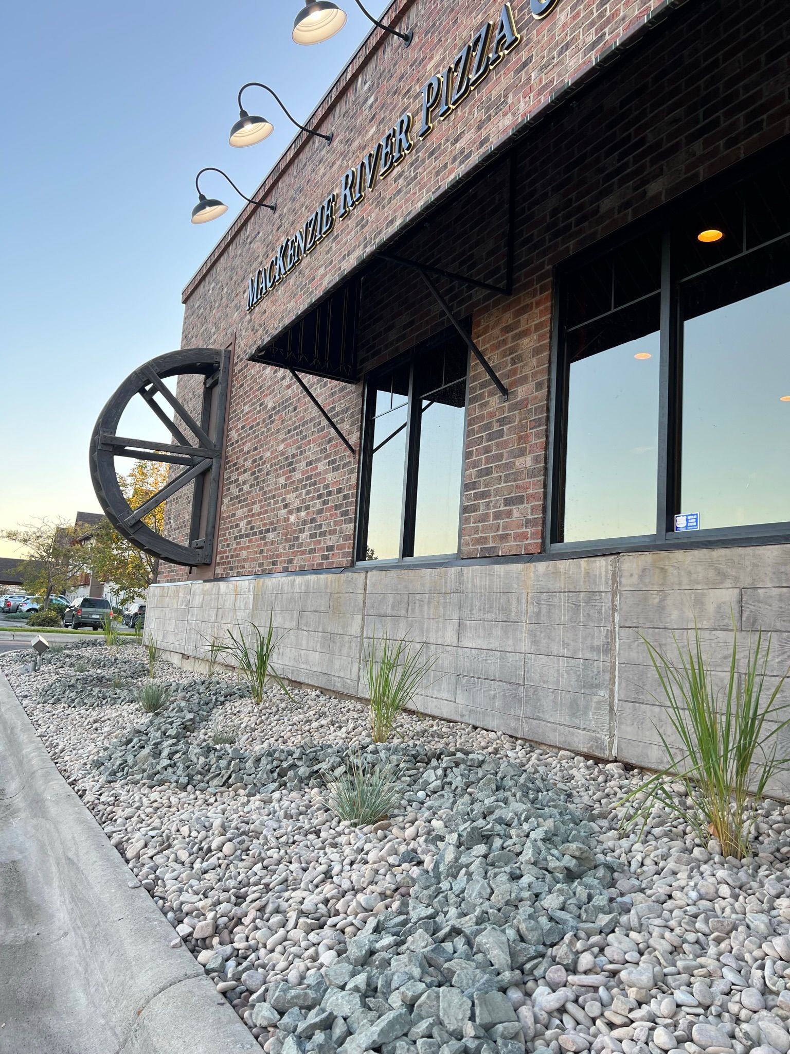 Exterior of a brick building with a large wheel, storefront windows, and landscaping.