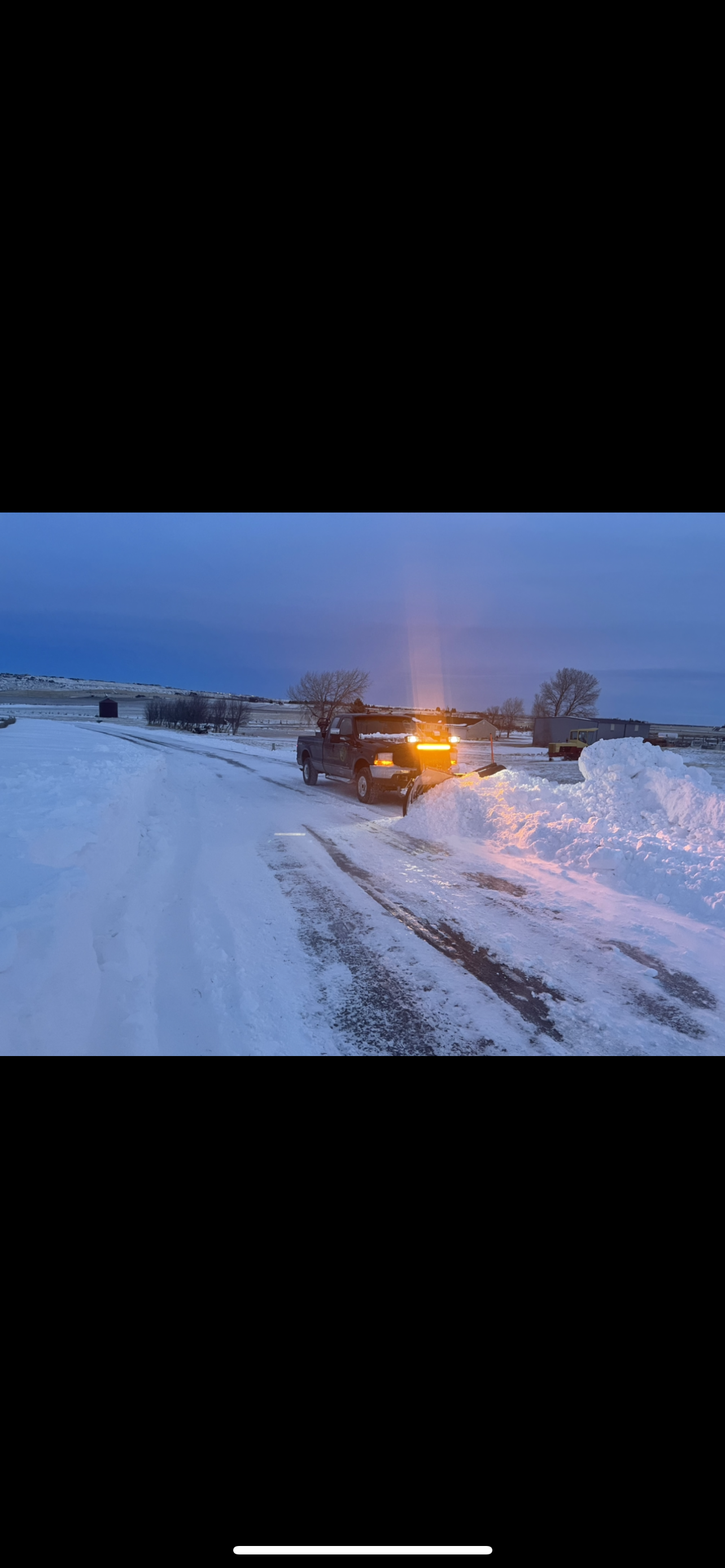 A snow-covered road with a vehicle plowing snow, illuminated by headlights. Blue sky background.