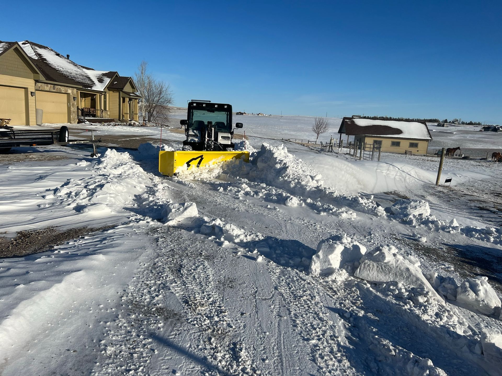 Yellow snowplow clearing a snowy driveway in a residential area on a sunny day.