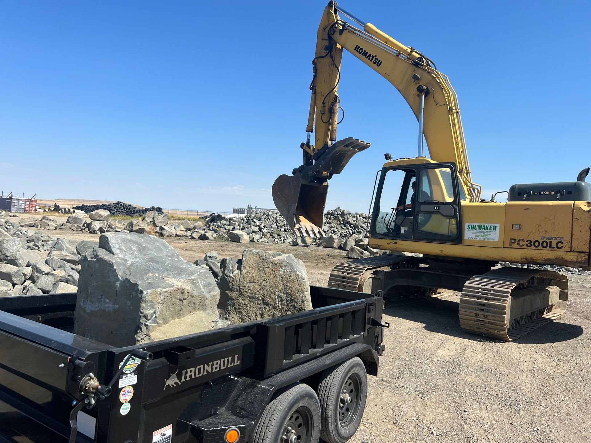Excavator loading large rocks into a black trailer on a sunny, outdoor worksite.
