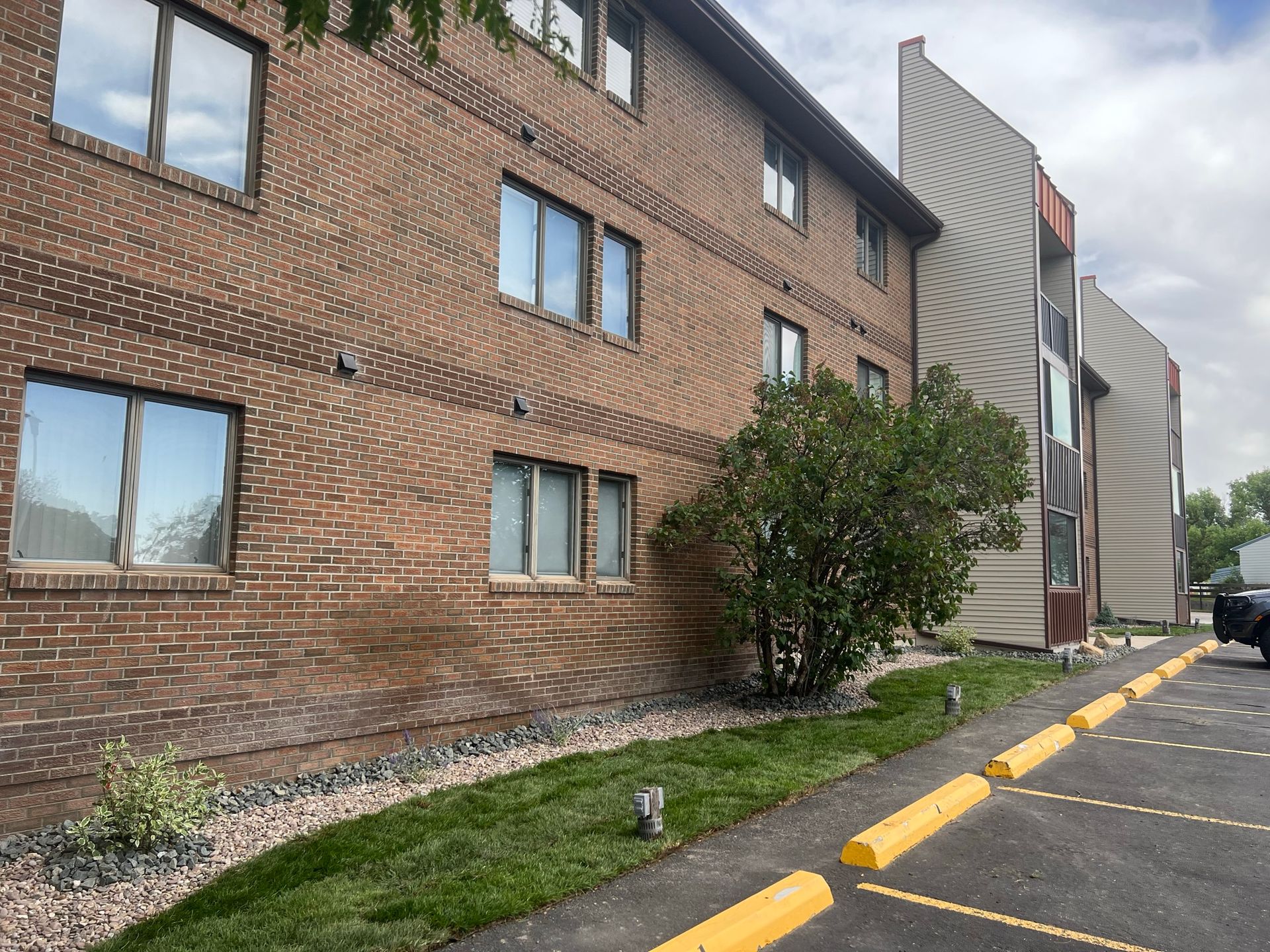 Brick building with windows, next to a parking lot, small lawn with a bush, and an angled wall with a different facade.