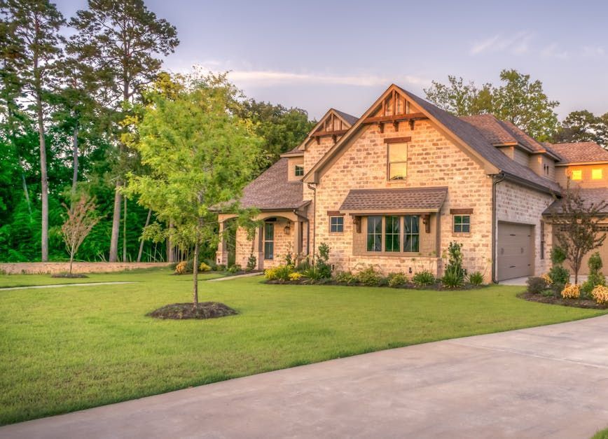 Stone house with a green lawn and driveway, surrounded by trees, during sunset.