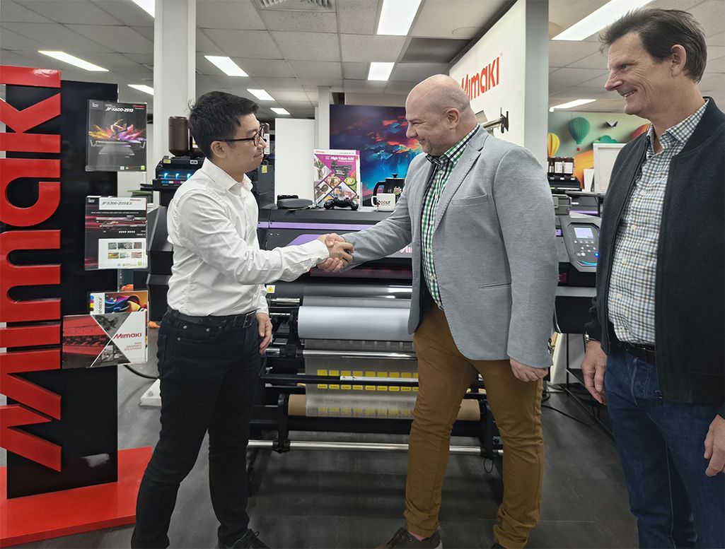 three men looking at each other, smiling and shaking hands in front of Mimaki