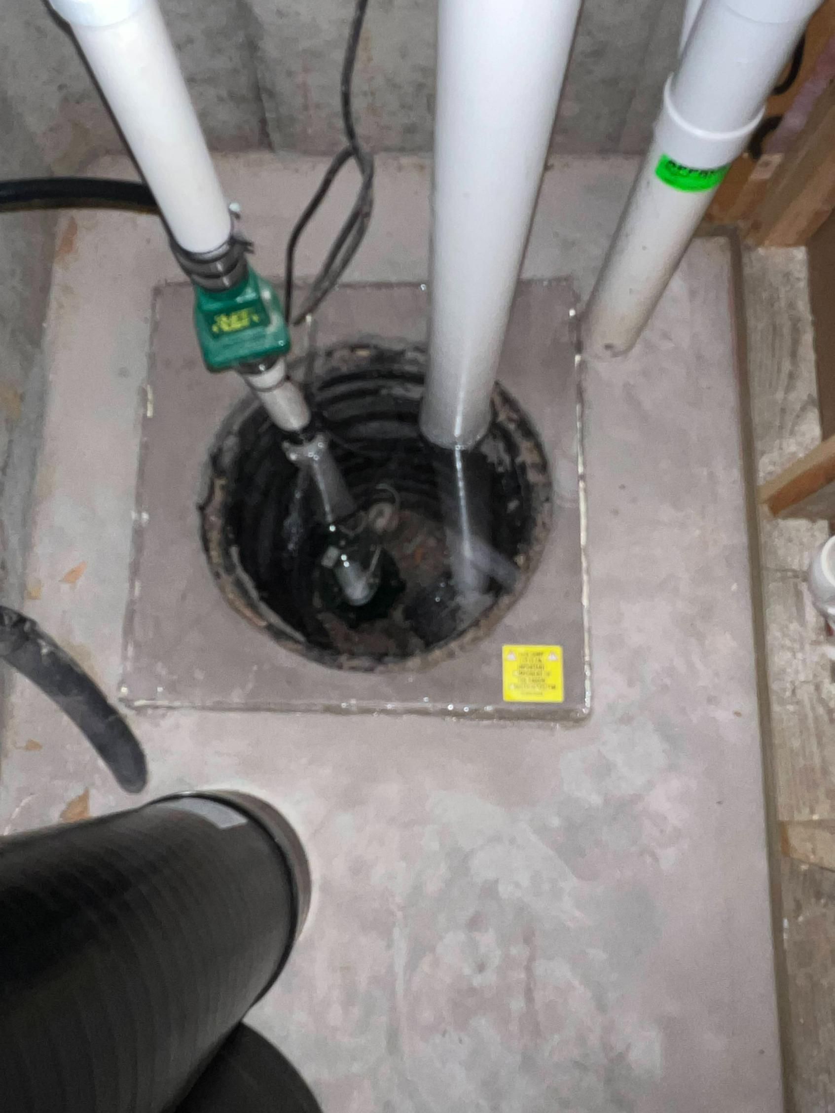 Overhead view of a sump pump inside a concrete pit with pipes attached; grey and white colors.