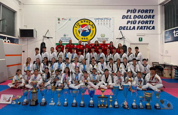 Foto di gruppo di una lezione di Taekwondo in uniforme bianca, in posa in palestra con uno striscione della scuola di Genova.