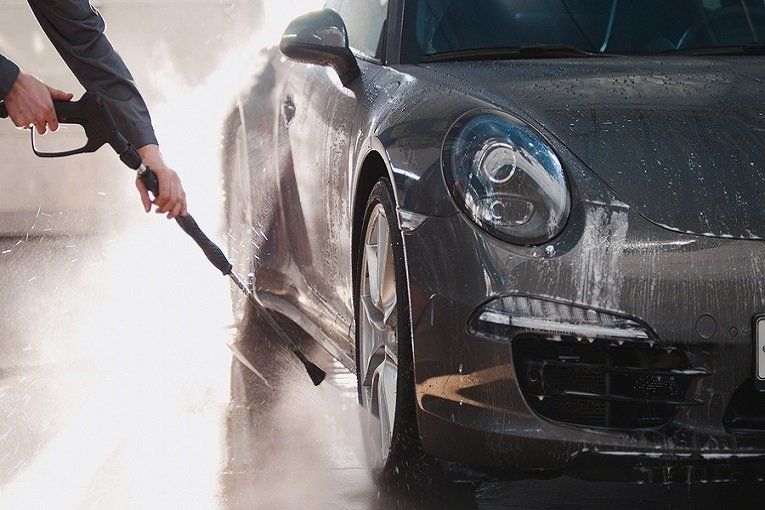 Person washing a gray sports car with a pressure washer in a car wash.