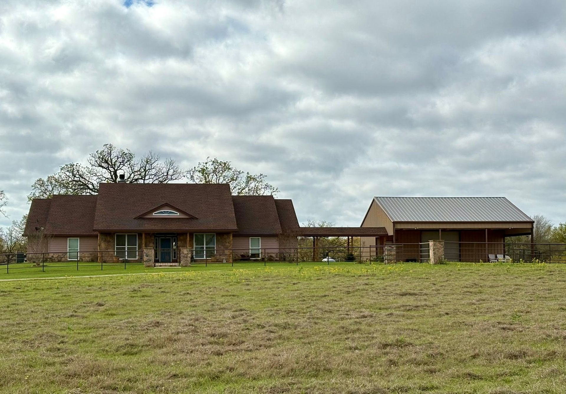 Rustic house with brown roof and detached garage in a grassy field under cloudy sky