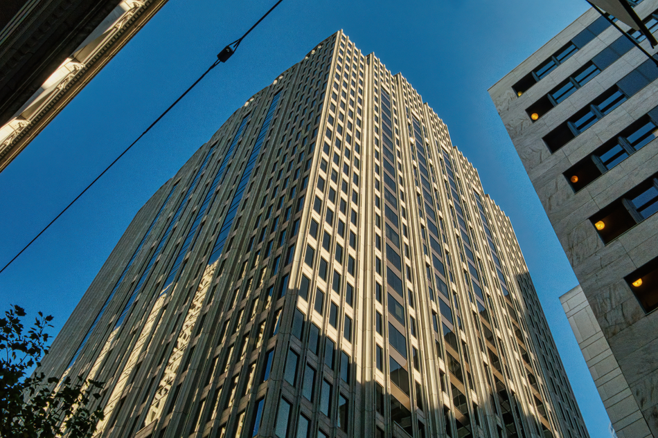 Tall office skyscraper seen from below against a clear blue sky, with neighboring buildings at the edges