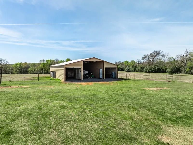 Ranch-style barn with open stalls in a fenced grassy field under a clear sky