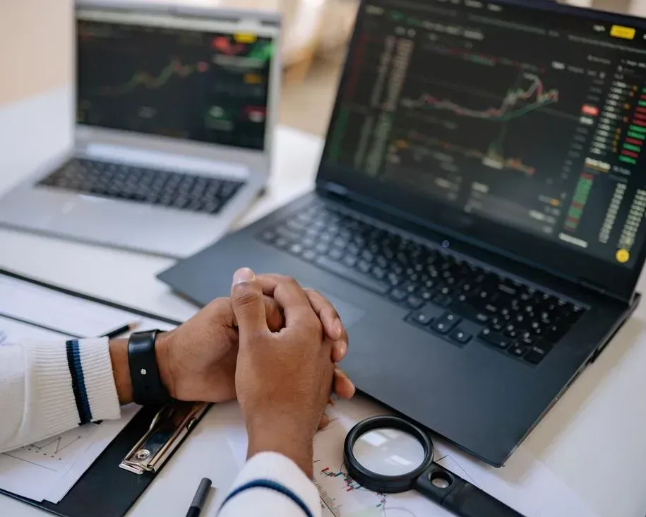 Hands clasped beside laptops showing stock charts on a desk with a magnifying glass