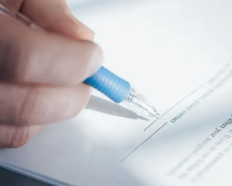 Hand holding a blue pen signing a document on a white desk