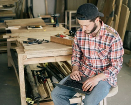 Man in plaid shirt working on a laptop at a wooden workbench in a workshop