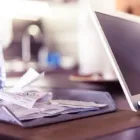 Laptop beside a tray of stacked receipts on a wooden desk in a blurred office setting