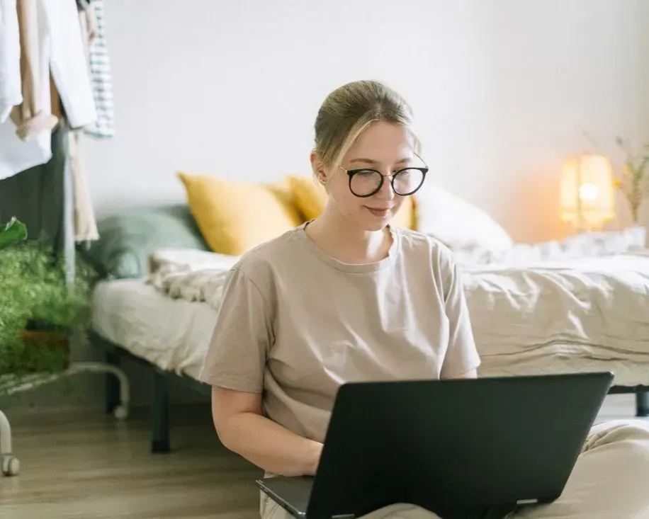 Person using a laptop in a bright bedroom with a bed and warm lamp in the background