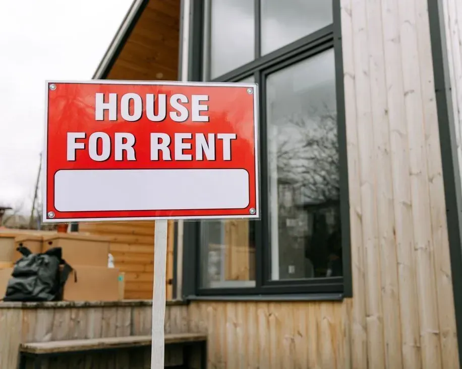 Red and white "House for Rent" sign outside a building with wood siding and windows