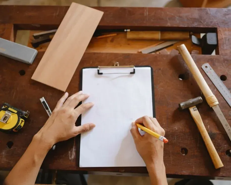 Hands sketching on a clipboard at a woodworking workbench with tools and wood pieces nearby