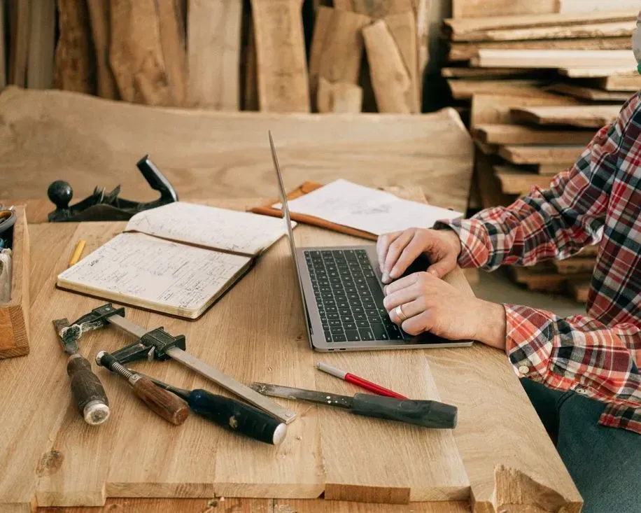 Person typing on a laptop at a wooden workbench with woodworking tools and an open notebook nearby