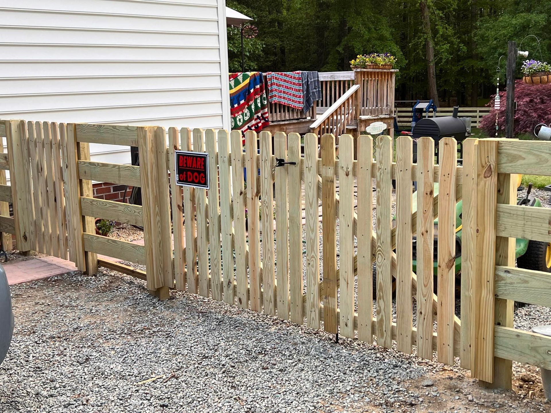 Wooden picket gate in a gravel yard beside a house, with a black and red sign attached to the fence.