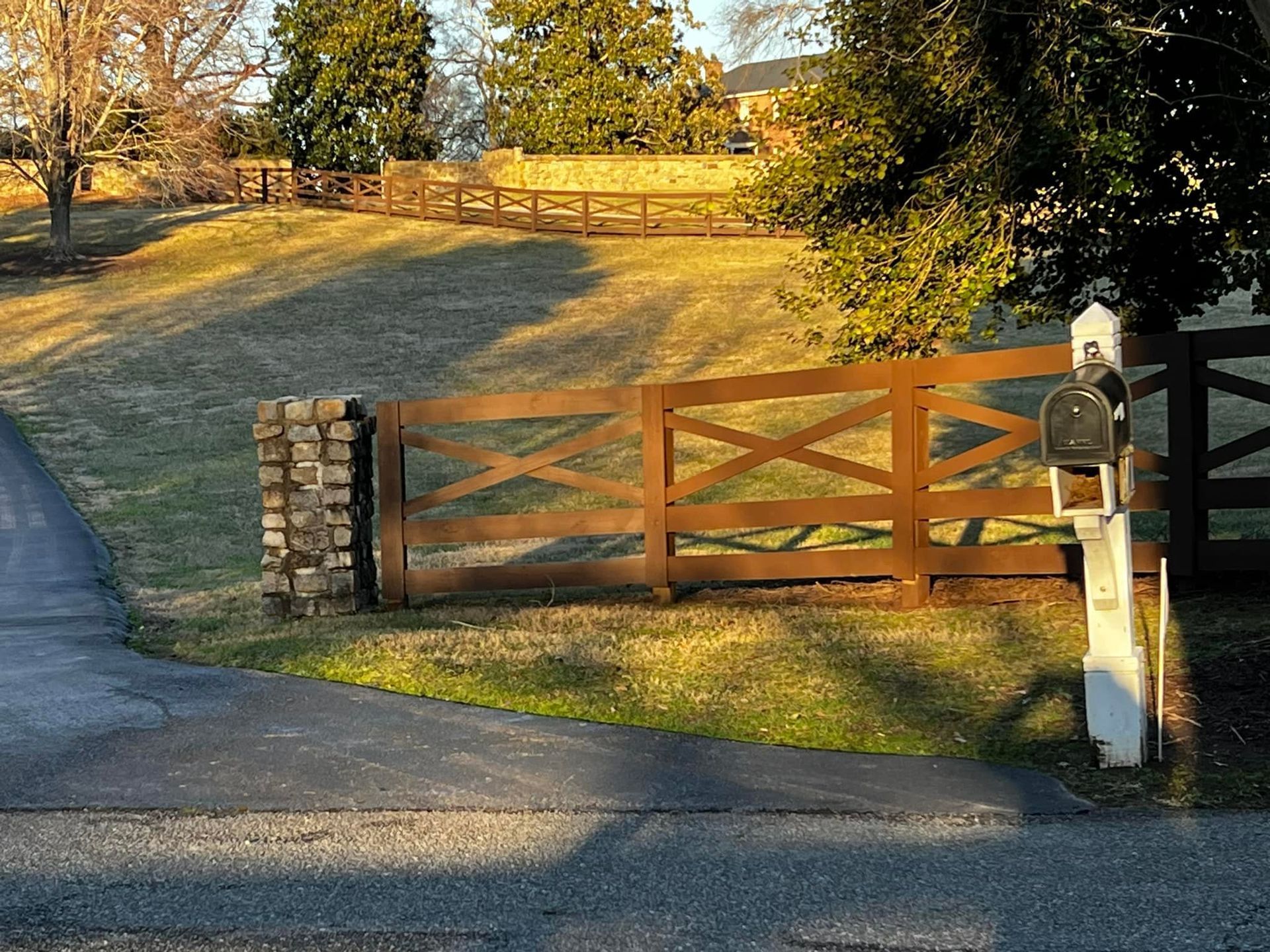 Rural driveway with open wooden gate, stone posts, and a mailbox at the entrance at sunset