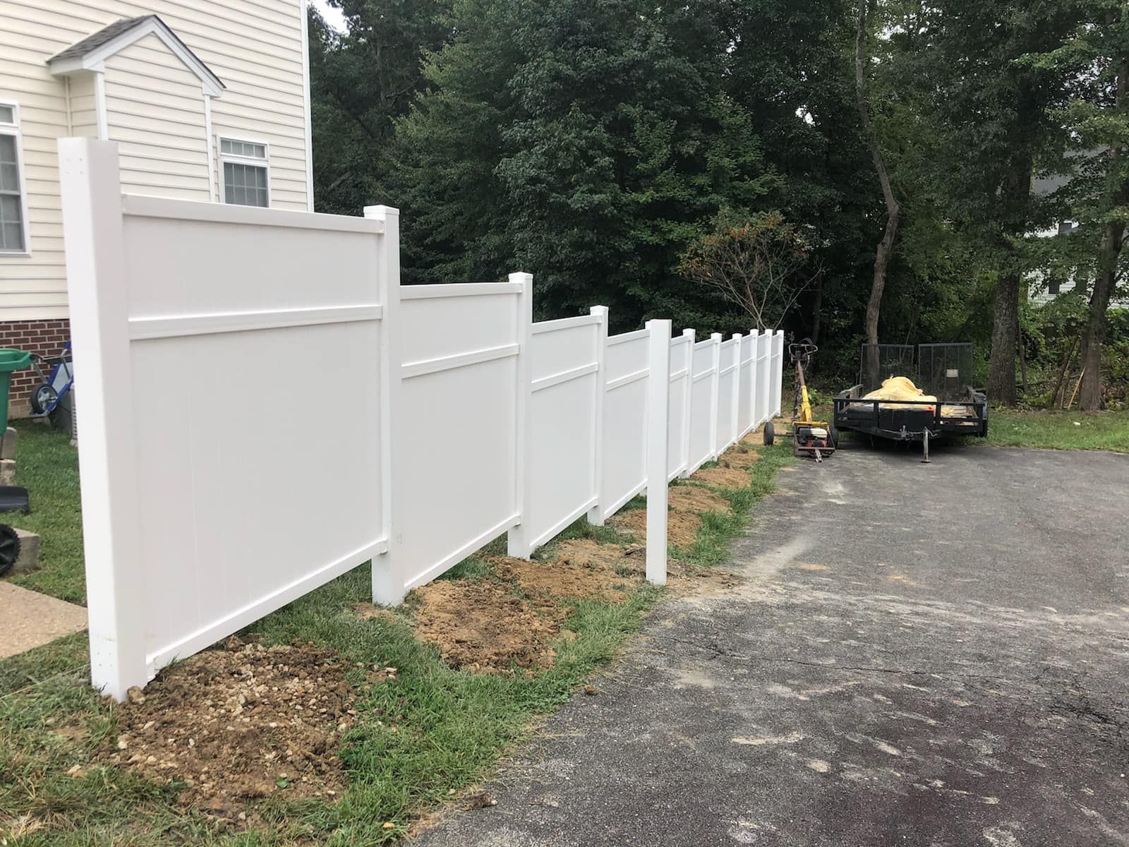 White vinyl fence along a driveway beside a house, with a lawn, trees, and a parked lawn tractor in the background
