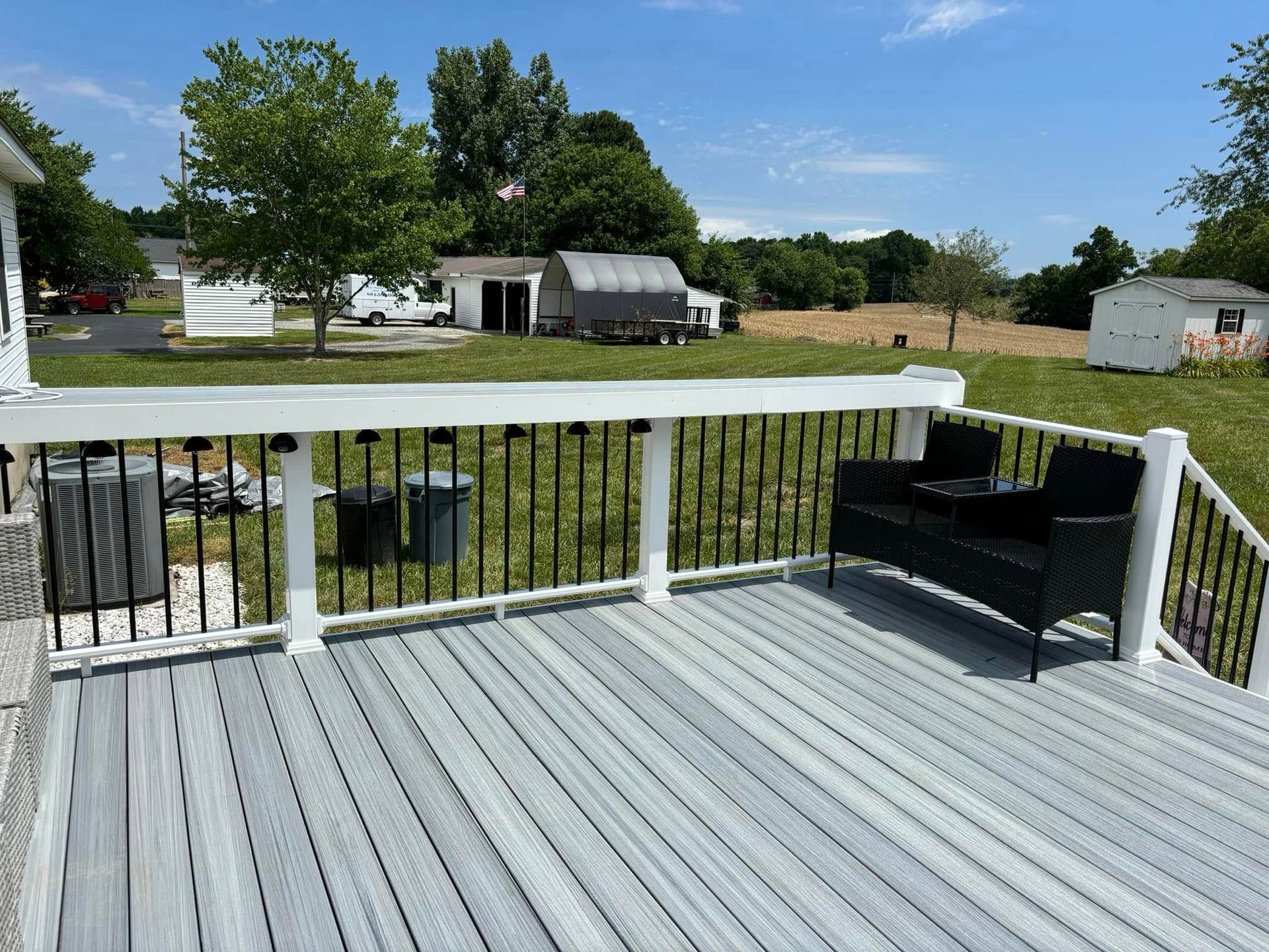 White deck overlooking a grassy yard with a black bench and rural outbuildings under a blue sky