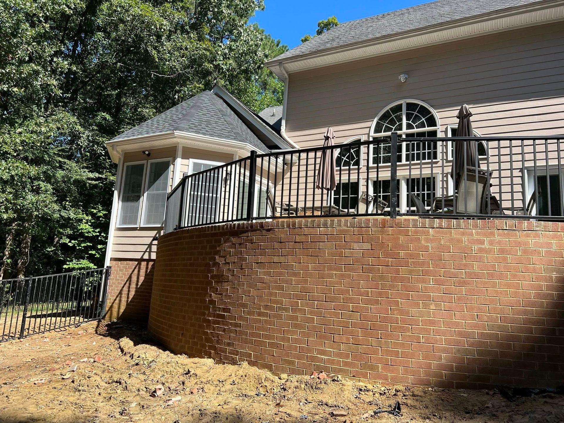 Brick patio wall and deck with black railings on a house, beside a small white gazebo in a wooded yard.