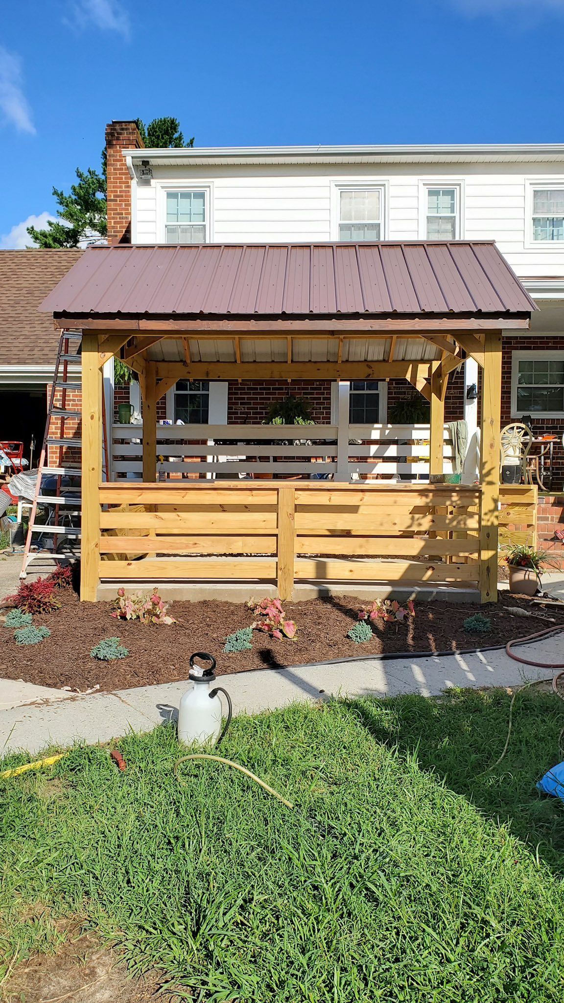 Wooden gazebo with a brown metal roof in a backyard garden, with a house in the background.