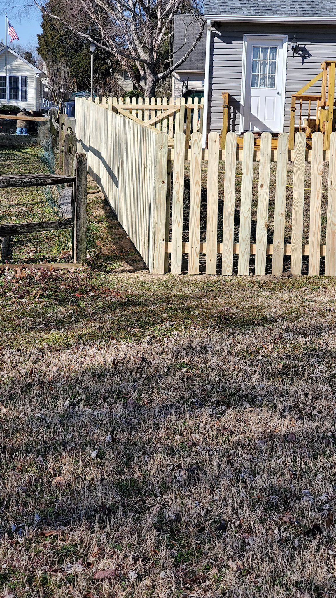 Weathered wooden fence around a small yard beside a house, with a wire fence on the left and fallen leaves in front.