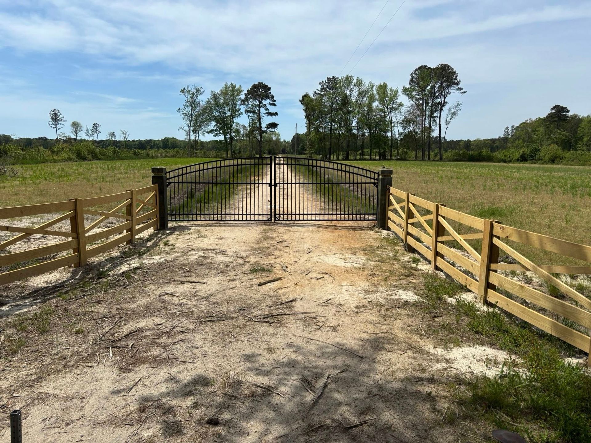 Gravel driveway leading to a black gate between wooden fences across a grassy field under a blue sky