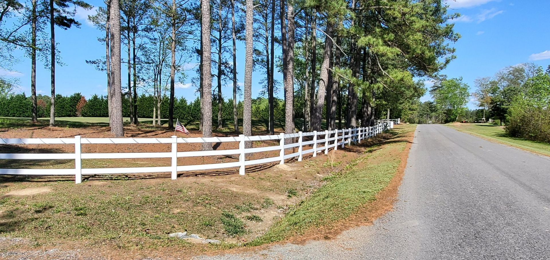 White fence along a sunny roadside beside pine trees and a gravel road