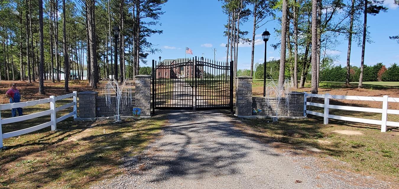 Gated driveway entrance with white fences and tall pine trees on a sunny day