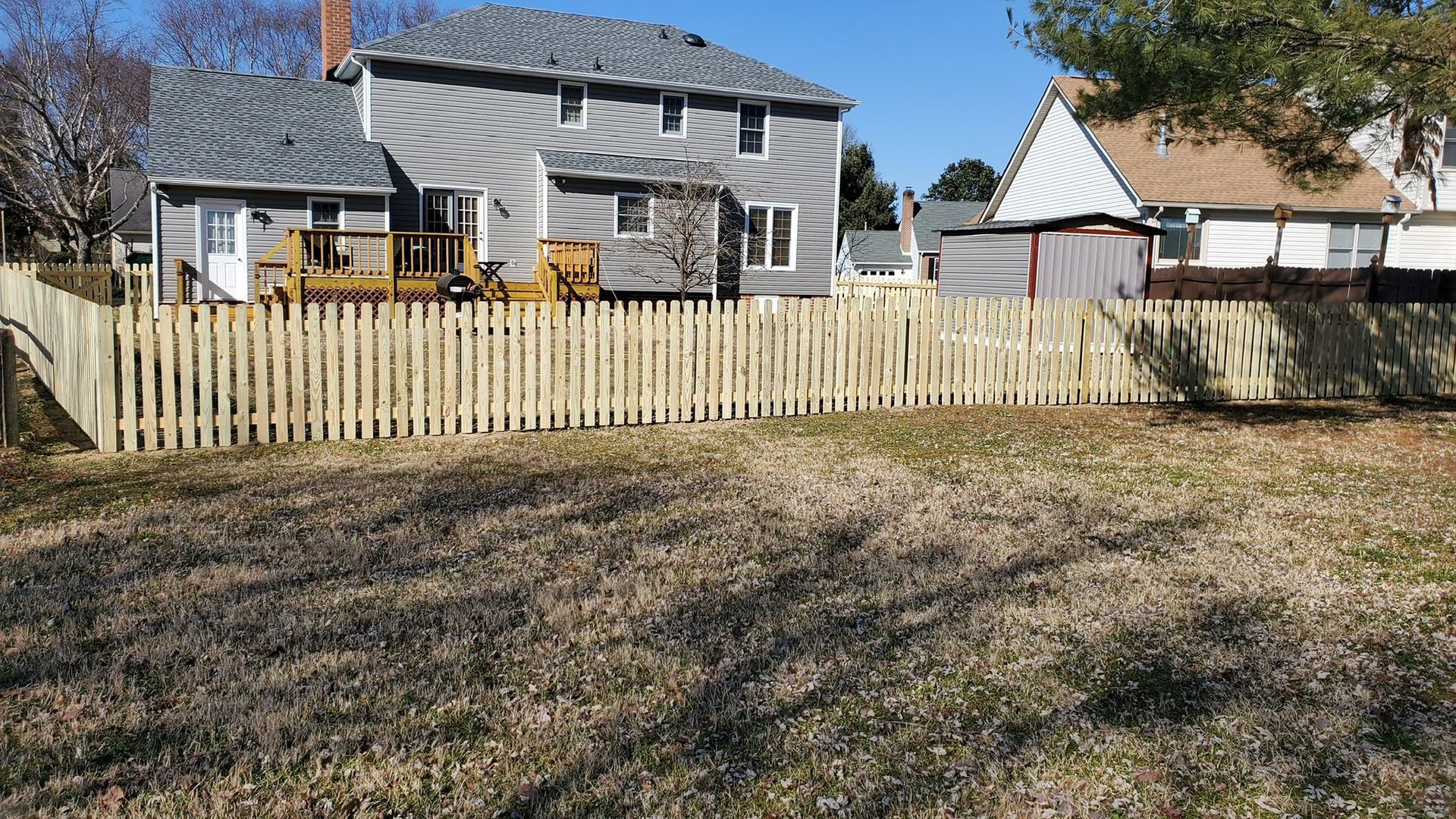 Backyard with a wooden fence, dry grass, and two houses under a clear blue sky