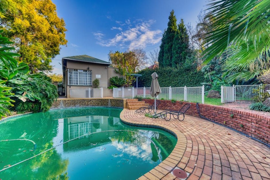 Backyard pool with curved brick patio, lounge chairs, white fence, and lush green trees under a blue sky