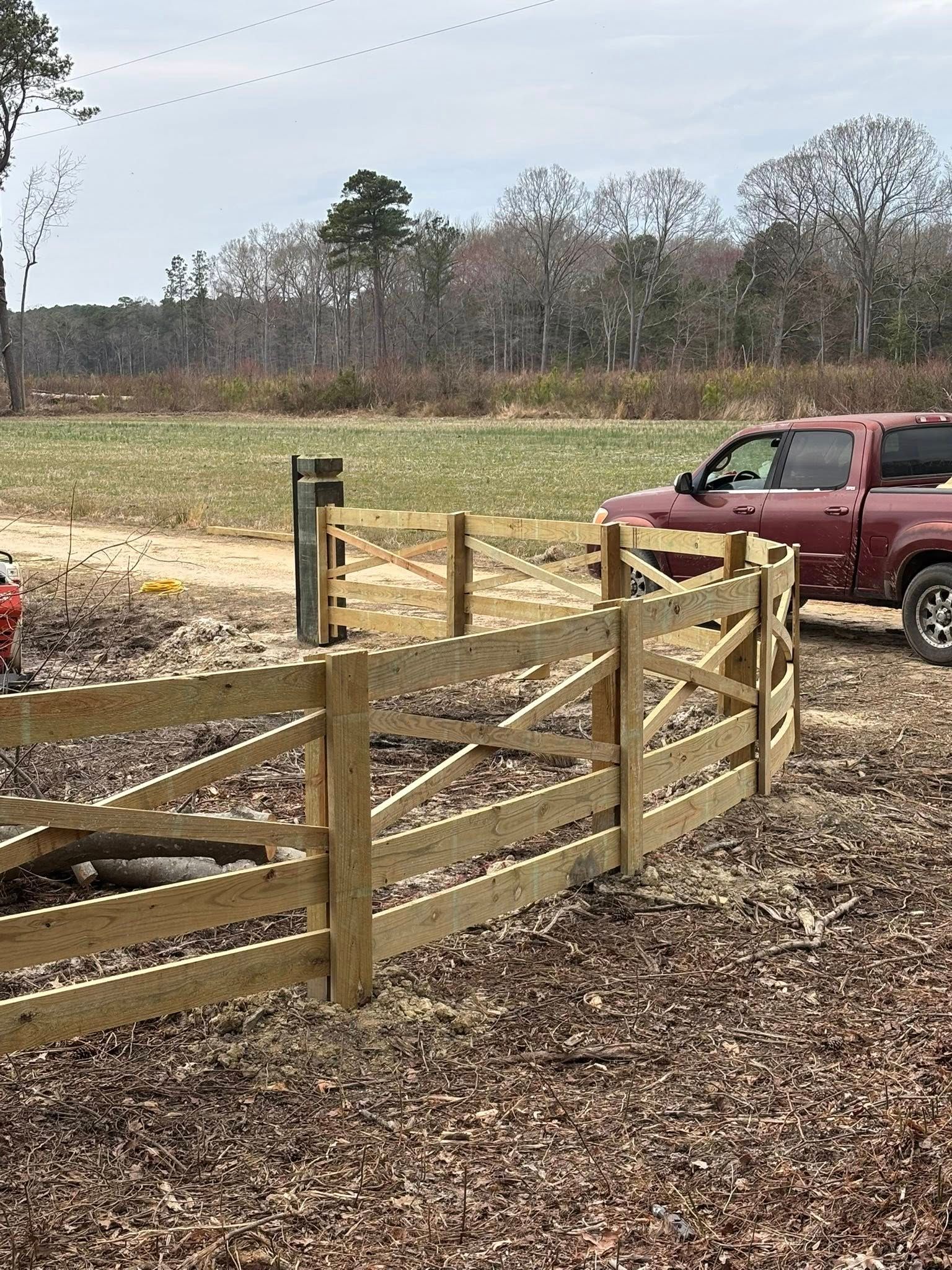 Wooden farm gate by a gravel driveway, with a red pickup truck parked beside a grassy field.
