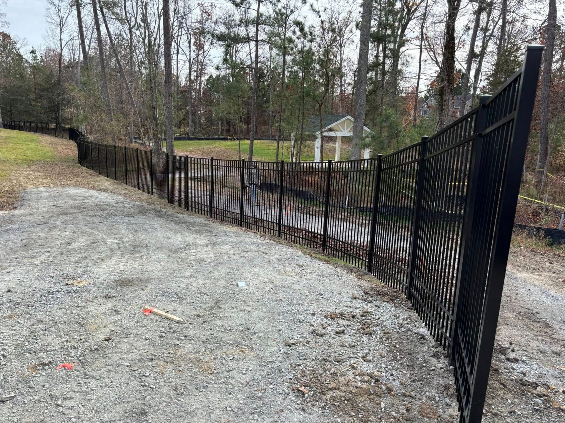 Black metal fence along a gravel path in a wooded yard, with houses in the background.