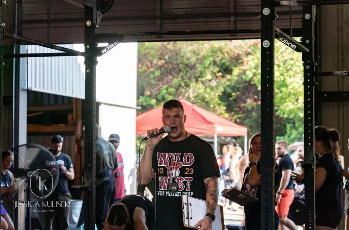 Man speaking on stage in a gym, with a crowd and red canopy visible behind him.