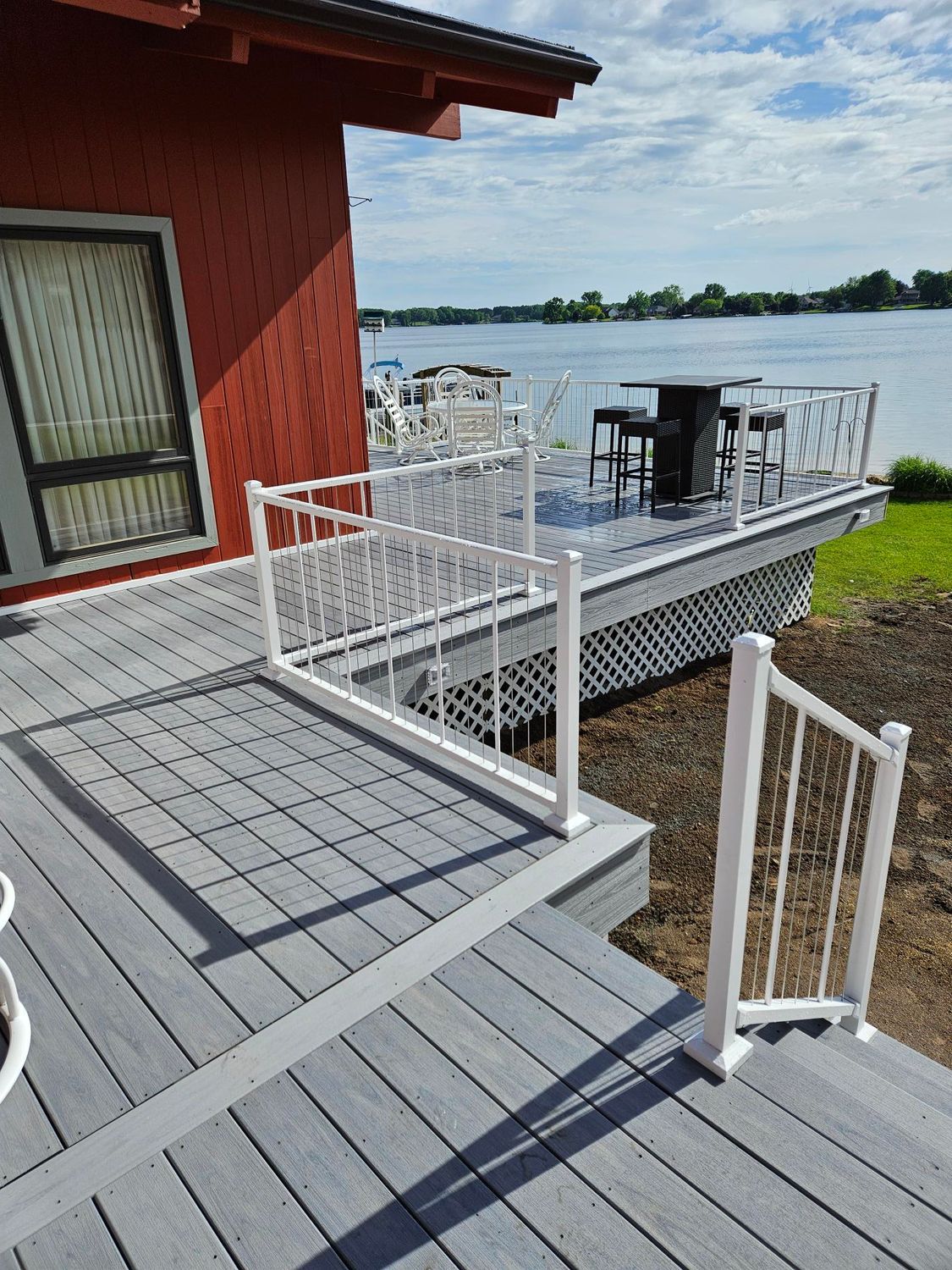Outdoor deck with white railings beside a red house, overlooking water with a table and chairs.