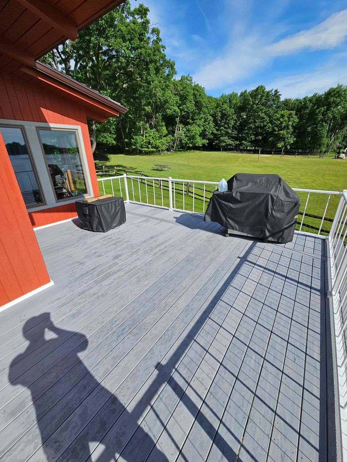 Red house deck with railing, grill, and open grassy yard under a sunny blue sky