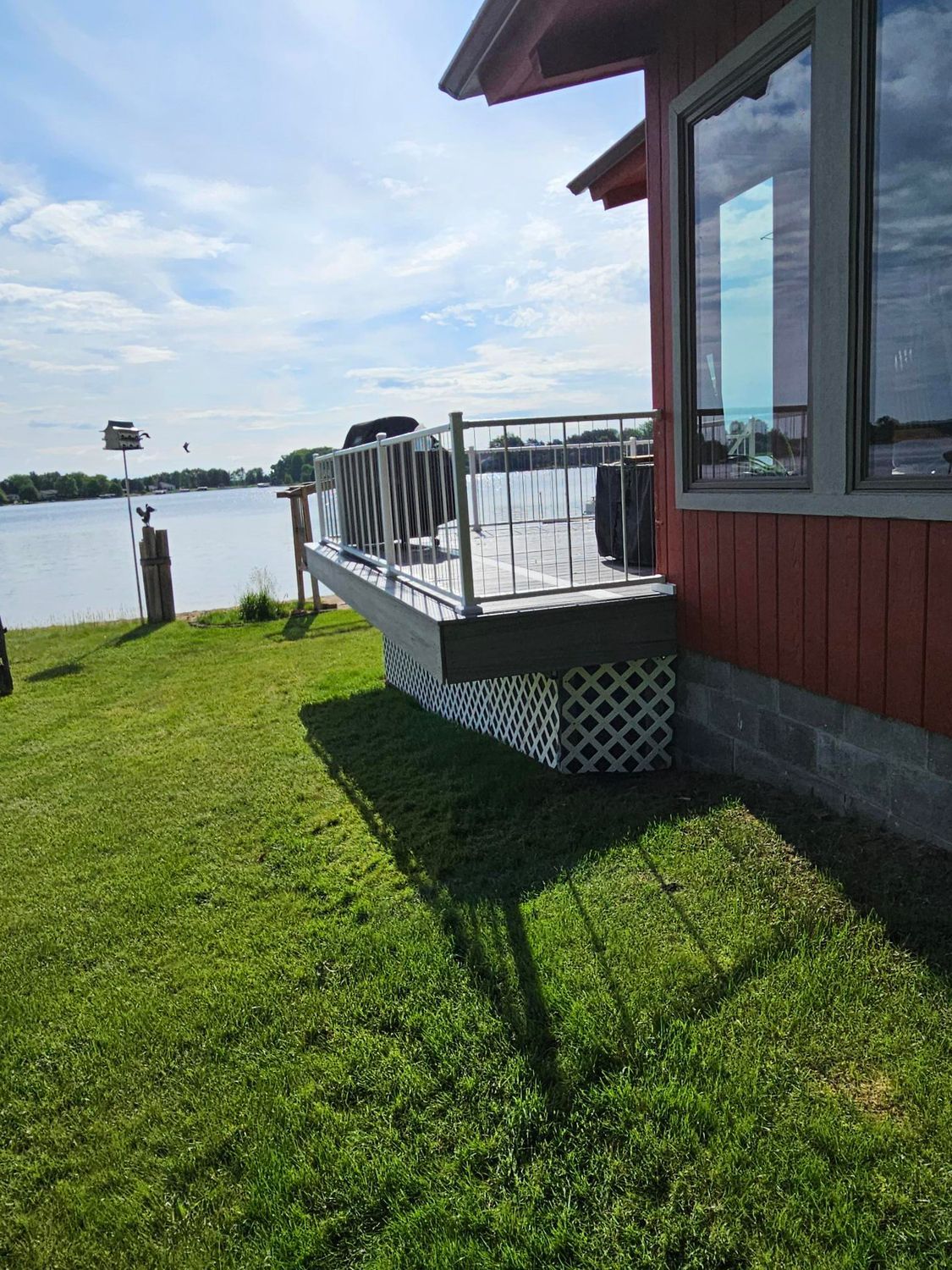 Red house porch with railing beside a grassy lakeside yard under a bright blue sky