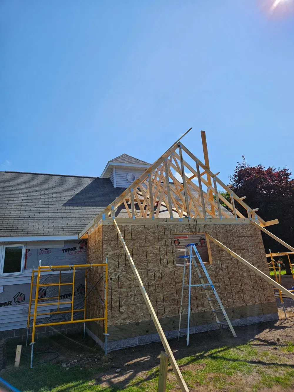 House under construction with exposed roof framing, ladders, and scaffolding under a clear blue sky