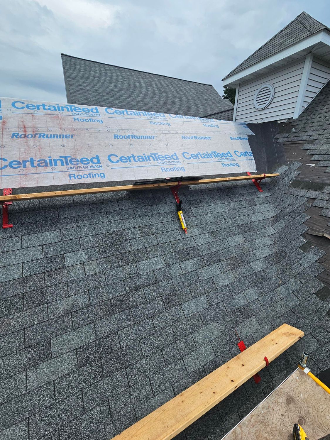 Roof under repair with gray shingles, underlayment, and a few wooden planks visible near a dormer window.