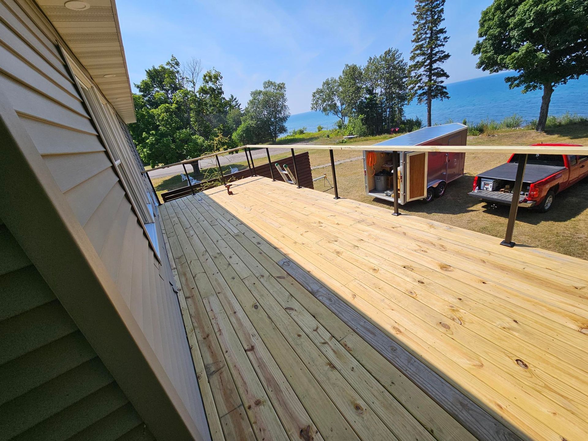 Wide wooden deck beside a house, overlooking a driveway, garage, and parked red car under a blue sky.