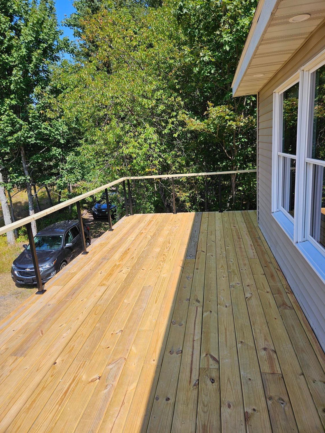 Wooden deck with railing beside a house, overlooking trees and parked cars